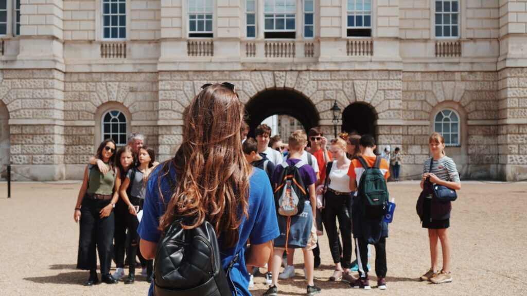 Group of international students gathered with a guide wearing a blue shirt in front of a large historic stone building with arches and windows.