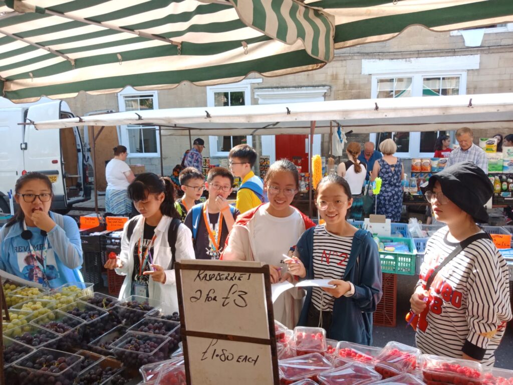 Group of students browsing fruit stalls at an outdoor market during a camp excursion