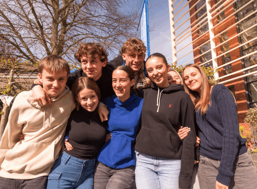 Smiling group of teenage students posing outdoors on a sunny day in front of a flag and modern school building.