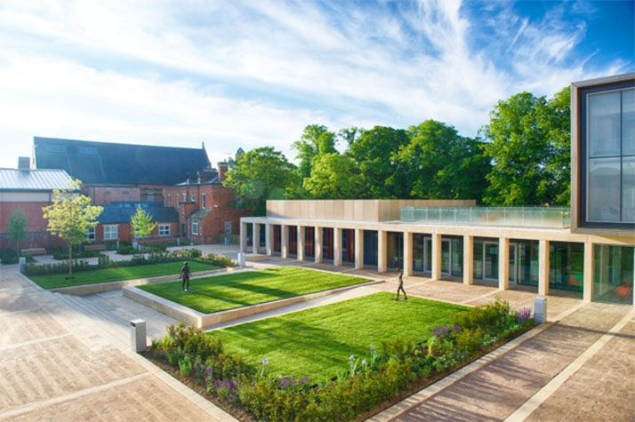 Modern courtyard with landscaped lawns and a colonnaded building, bordered by traditional school architecture and trees.